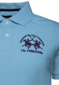 Ljusblå polo skjorta i strukturerad tyg. Har en lila broderad logotyp av två polospelare och namnet "LA MARTINA." Tre knappar på kragen.