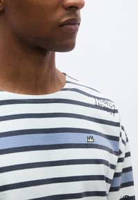 Ριγέ μπλούζα με μακριά μανίκια σε μπλε, ναυτικό και λευκό. Διαθέτει μικρό κίτρινο λογότυπο κορώνας και τη λέξη "RODEO" κεντημένη στον ώμο. Απαλό ύφασμα.