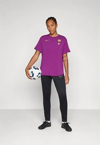 Purppura Nike -t-paita, jossa on Barcelonan logo, ja mustat urheiluhousut, joita pidetään jalkapallon kanssa, rento istuvuus ja sileä pinta.
