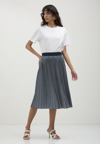 Donna con una maglietta bianca a maniche corte, una gonna midi plissettata a zigzag blu e bianca, e sandali bianchi con tacco, che posa contro uno sfondo chiaro e semplice.