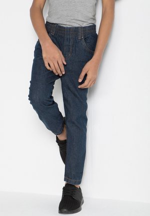 Pantalon en denim avec une taille élastique, légèrement effilé aux chevilles, de couleur bleu foncé, avec des coutures contrastantes. Des baskets noires complètent le look.