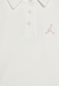 Vit polo skjorta med strukturerad tyg, med krage och tre knappar. Anmärkningsvärd rosa broderad Jumpman-logotyp på vänster sida.
