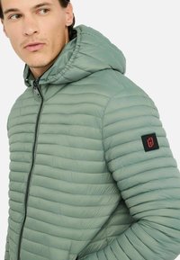 Lys grøn pudejakke med hætte, der har horisontalt syede sektioner og et sort logopatch på ærmet. Lynlåslukning foran.