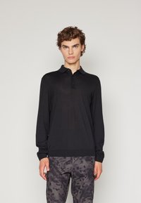 Camisa polo de manga longa preta com um fecho de botões e bainha canelada, combinada com calças cinzas estampadas, apresentando uma textura suave.