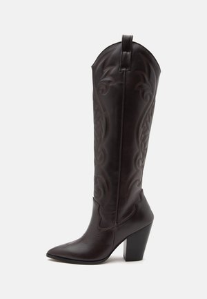 Botte de cowboy haute en cuir marron foncé avec coutures décoratives, bout pointu et talon haut épais et angulé sur fond blanc.
