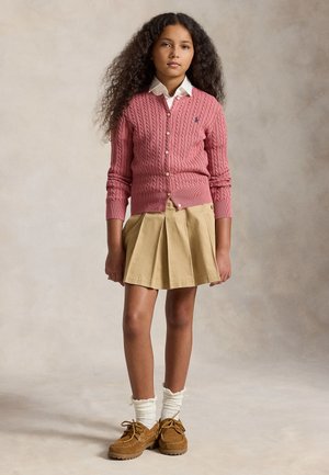 Pige med krøllet hår iført en lyserød kabelstrikket cardigan, hvid skjorte med krave, beige plisseret nederdel, hvide sokker og brune sko, stående mod en ensfarvet baggrund.