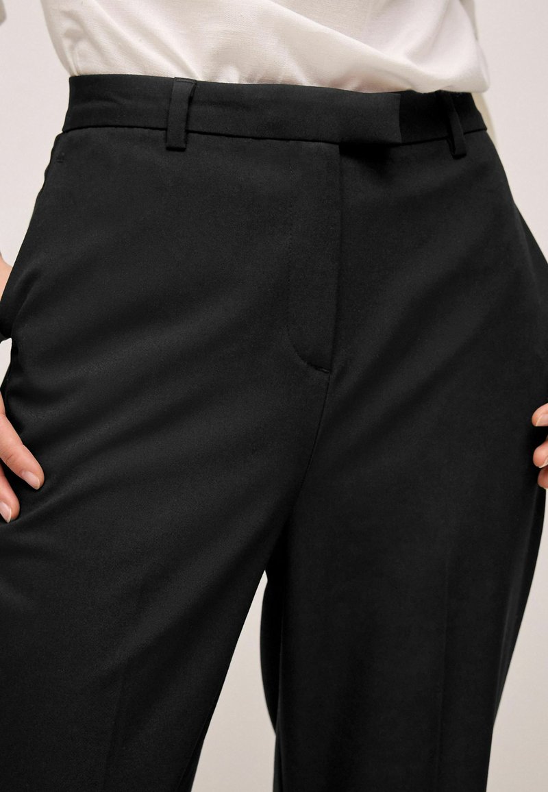 Musta räätälöity housut, joissa on sileä pinta, slim fit -leikkaus ja etutaskut, tyylitelty suoran leikkauksen ja minimaalisten yksityiskohtien kanssa.