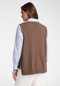 Gilet di maglia marrone con orlo e giromanica a costine, indossato sopra una camicia azzurra chiara. Presenta un piccolo logo sul retro del collo.