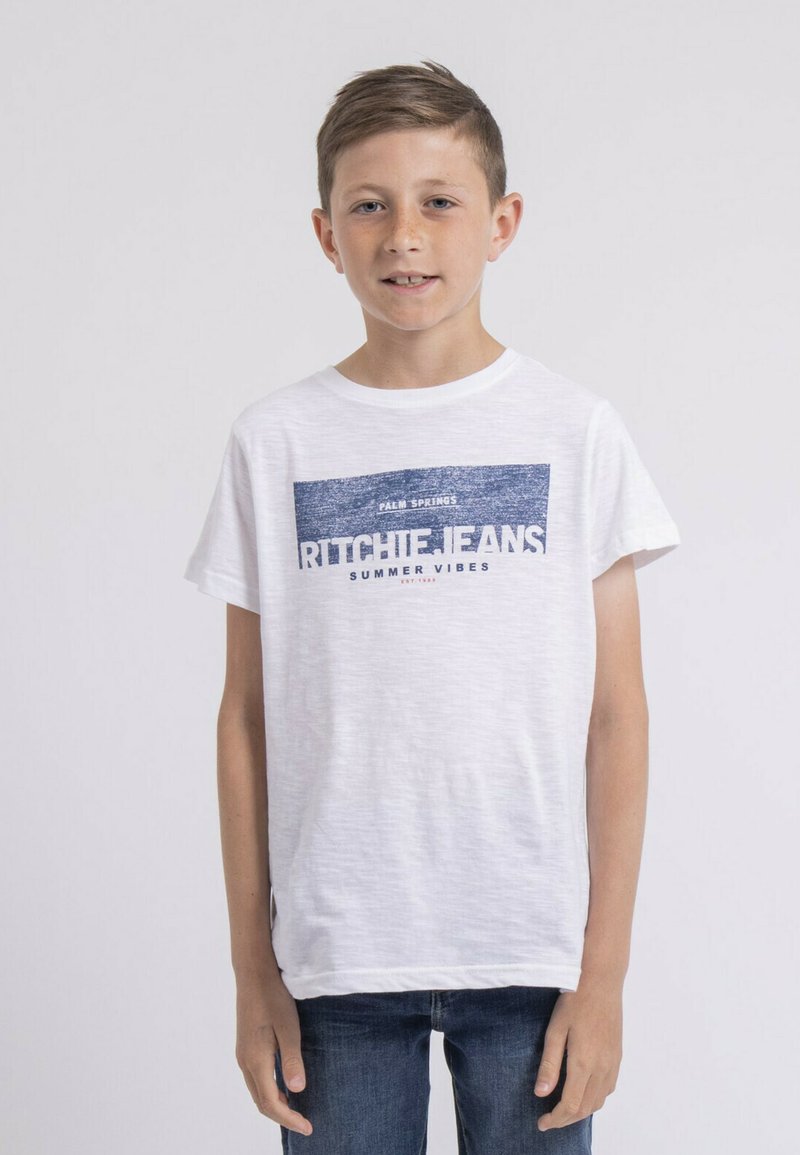 T-shirt en coton blanc à manches courtes présentant un design graphique bleu incluant du texte et des rayures. Porté avec un jean en denim bleu.