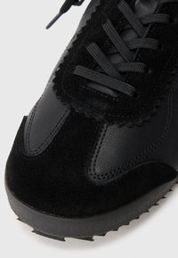 Schwarzer Sneaker aus einer Mischung von glattem Leder und Wildleder, mit strukturierten Akzenten, flachen schwarzen Schnürsenkeln und einer robusten Gummisohle.
