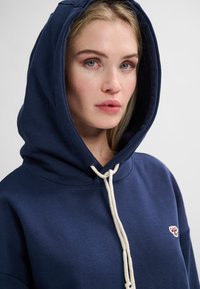 Mörkblå huvtröja med dragsko och en liten broderad logotyp. Mjukt material, avslappnad design och stor huva för skydd.