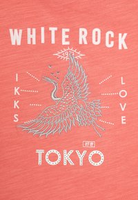 Koraļļu kokvilnas T-krekls ar baltu un sudraba izšuvumu, kurā redzams gūbja dizains, teksts "WHITE ROCK," "TOKYO" un dekoratīvi akcenti.