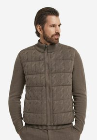 Braune Weste mit Reißverschluss, die vorne ein Zopfmuster und an den Ärmeln sowie am Saum Rippenstrick aufweist, hergestellt aus einem weichen, strukturierten Stoff.