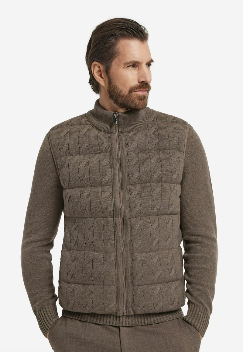 Braune Weste mit Reißverschluss, die vorne ein Zopfmuster und an den Ärmeln sowie am Saum Rippenstrick aufweist, hergestellt aus einem weichen, strukturierten Stoff.