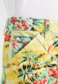 Gul denimtyg med tropiskt blommönster i rött, rosa och grönt. Inkluderar metallaccenter och ett synligt varumärkesetikett.