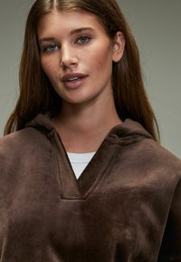 Brauner Cord-Hoodie mit V-Ausschnitt. Weicher Griff und lässige Passform. Weißes Unterhemd ist am Ausschnitt sichtbar.