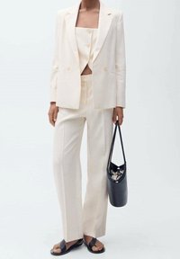 Veste beige cintrée à double boutonnage et pantalon à jambes larges, assortis à un crop top et un sac à bandoulière noir, portés avec des sandales plates.