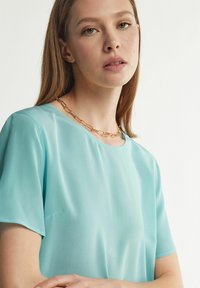 Ljusblå kortärmad blus i genomskinligt tyg med rund halsringning, har en slät textur och lagerdesign. Accentuerad med ett guldhalsband.