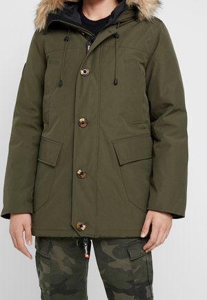 Mężczyzna ubrany w oliwkowo-zieloną parkę z kapturem wykończonym futrem i zatrzaskami, w połączeniu z zielono-czarnymi bojówkami moro.