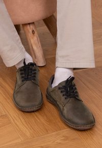 Dunkelolivgrüne Wildlederschuhe mit schwarzen Schnürsenkeln, runder Spitze und heller Naht, getragen über weißen Socken, auf einem Holzboden positioniert.