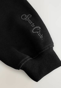 Sort sweatshirtærme med ribbet tekstur. Har et broderet gråt logo, "Sports Club," nær enden. Glat stofoverflade.