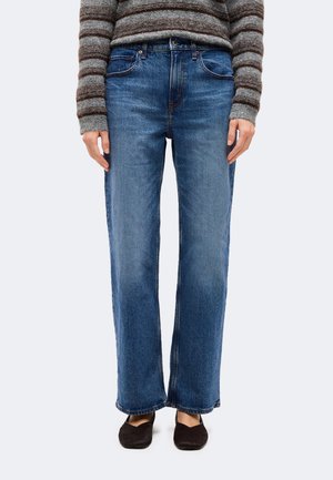 Personne portant un jean bleu droit, un pull rayé marron et gris, et des chaussures plates noires, debout devant un fond uni.