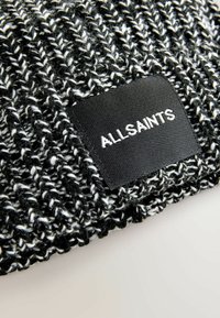 Črna in bela pletena kapa z teksturiranim vzorcem. Ima pravokoten črn oznako z napisom "ALLSAINTS" v belem.
