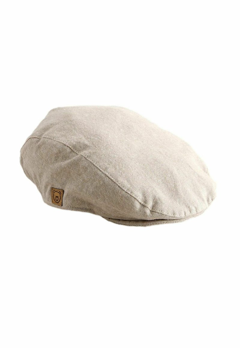 Boina plana beige claro hecha de tela suave, con una forma redondeada y un pequeño parche de cuero con un logo de oso en el lateral.