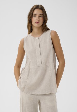 Jeune femme portant un top en lin beige sans manches et un pantalon large assorti, debout avec les mains dans les poches devant un fond uni.