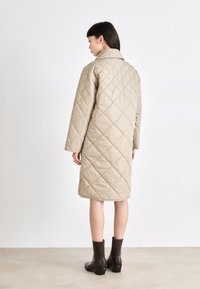 Manteau beige matelassé avec un col montant et des manches longues, présentant un motif en losanges et une coupe au niveau des genoux, associé à des bottines marron.