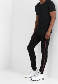 Jeans ajustados negros con una franja lateral roja, detalles texturizados, combinados con una camiseta negra sencilla y zapatillas deportivas blancas.