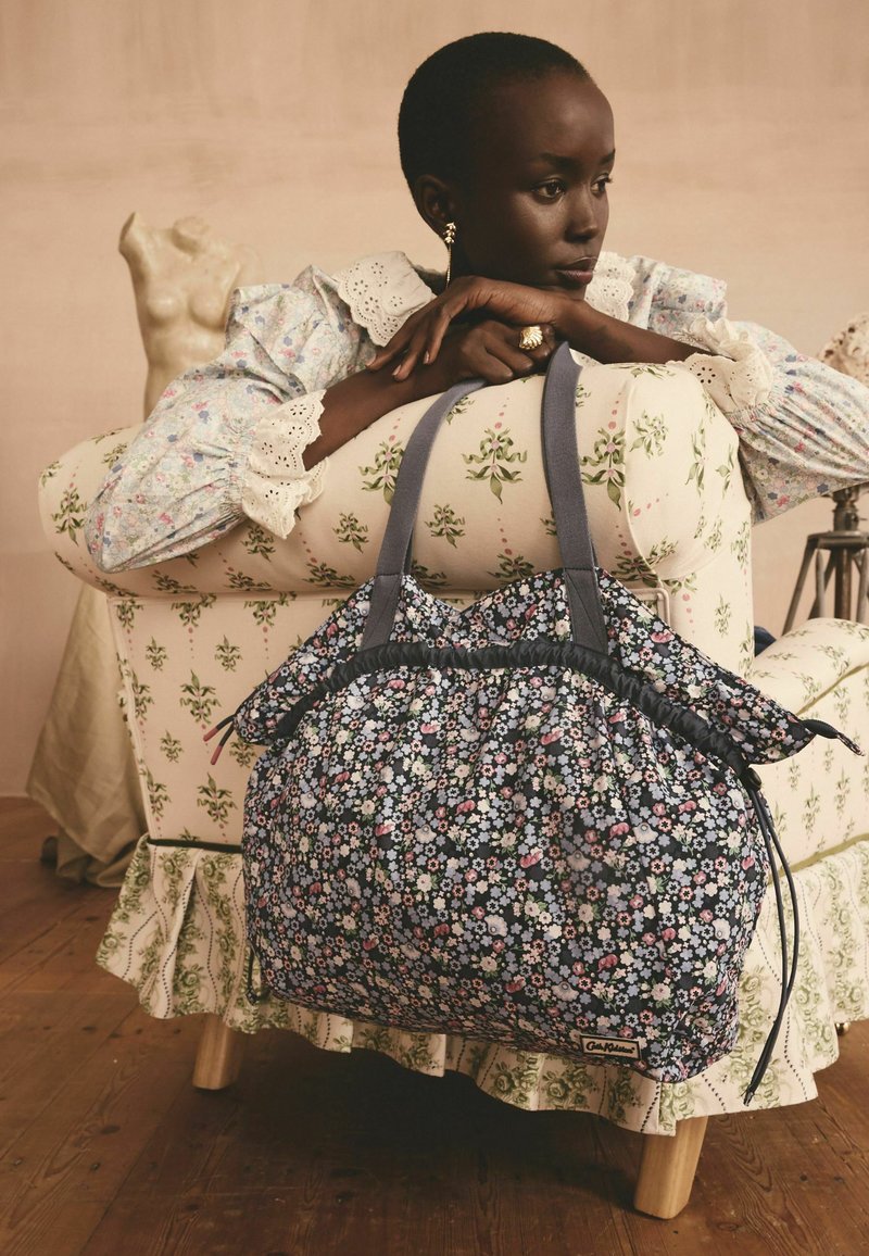 Sac fourre-tout floral avec un fond sombre, présentant un design froncé, des bretelles grises et une fermeture à cordon. Tissu doux avec une finition texturée.