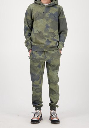Camo hoodie and pants in various shades of green, featuring a hood, text detail, and ribbed cuffs. Worn with colorful sneakers.
