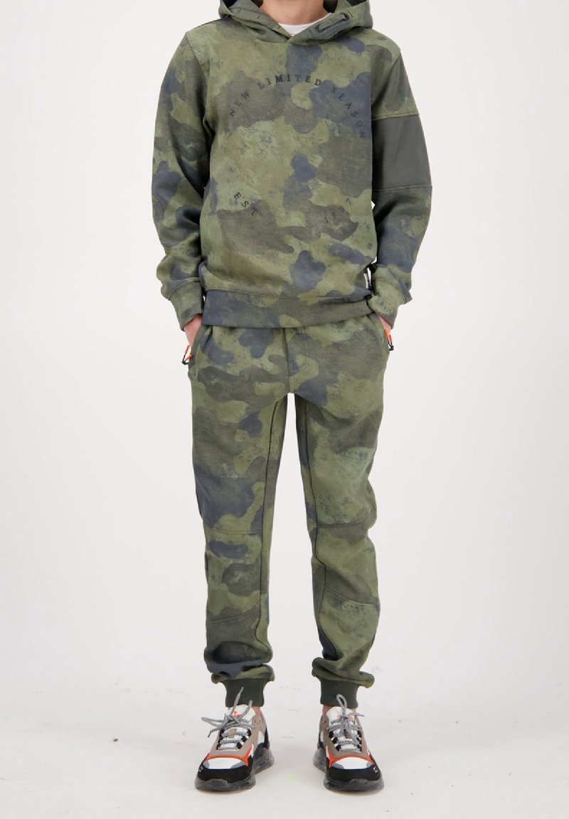 Camo hoodie og bukser i forskellige nuancer af grøn, med hætte, tekstdetalje og ribbede manchetter. Bærer farverige sneakers.
