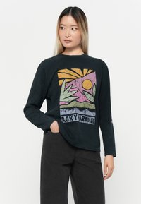 Svart langermet T-skjorte med et teksturert grafisk design som viser hender, sol og fjell i pastellfarger, merket "ROXY PARADISE."
