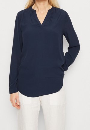 Blusa a maniche lunghe blu navy con scollatura a V e dettagli plissettati, realizzata in tessuto liscio. Orlo arrotondato ai lati, indossata con pantaloni in lino di colore chiaro.