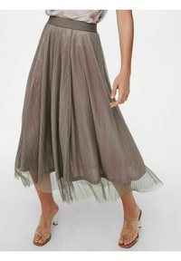 Jupe midi plissée en taupe avec une superposition transparente, portée avec des sandales à talons à straps transparents et un chemisier léger rentré.