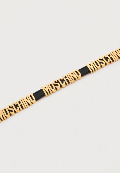 Χρυσά γράμματα που σχηματίζουν τη λέξη «MOSCHINO» επανειλημμένα σε μαύρο δερμάτινο λουράκι πάνω σε απλό λευκό φόντο.