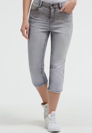 Woman wearing faded gray cropped jeans and white athletic shoes, standing with one hand in pocket and one hand relaxed.