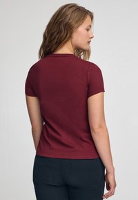 Camiseta de manga corta color burdeos con cuello redondo. Tela suave y diseño ajustado, vista desde atrás, combinada con pantalones oscuros.