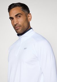 Balta sporta ceturtdaļas zip veste ar tumši zilām malām, kurai ir maza markas birka uz krūšu daļas. Gluda, viegla auduma.