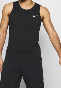 Mies, jolla on päällään musta hihaton Nike-paita ja mustat Nike-shortsit, seisoo yksinkertaista vaaleaa taustaa vasten.