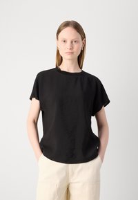 Haut noir à manches courtes en tissu léger et texturé avec un col rond. La coupe est décontractée, associé à un pantalon beige ample.