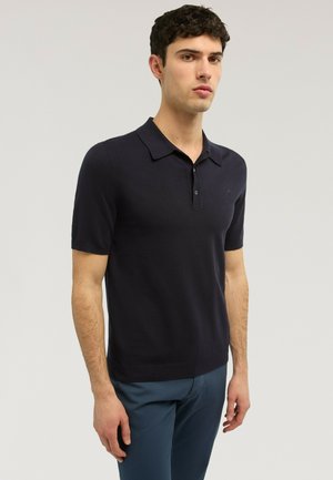 Joven con cabello corto y rizado, que lleva una camisa polo negra ajustada de manga corta y pantalones azules, posando contra un fondo claro.