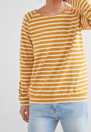 Personne portant une chemise à manches longues rayée jaune moutarde et blanche et un jean bleu clair, debout devant un fond clair uni.