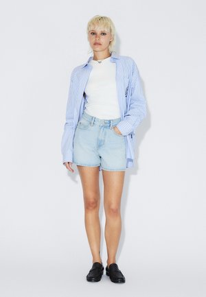 Camicia a righe blu a maniche lunghe sopra una t-shirt bianca, abbinata a pantaloncini di denim azzurro chiaro e mocassini neri. Outfit semplice e casual.