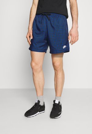 Mężczyzna ubrany w niebiesko-czarne szorty w kratę Nike, czarne sneakersy Nike, szare skarpetki i czarną koszulkę, stojący na jednolitym podłogu.