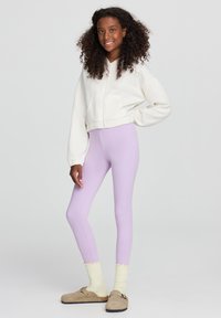 Leggings viola con una texture liscia, abbinati a un hoodie bianco corto e calzini giallo chiaro con bordo scallop, indossati con zoccoli beige.