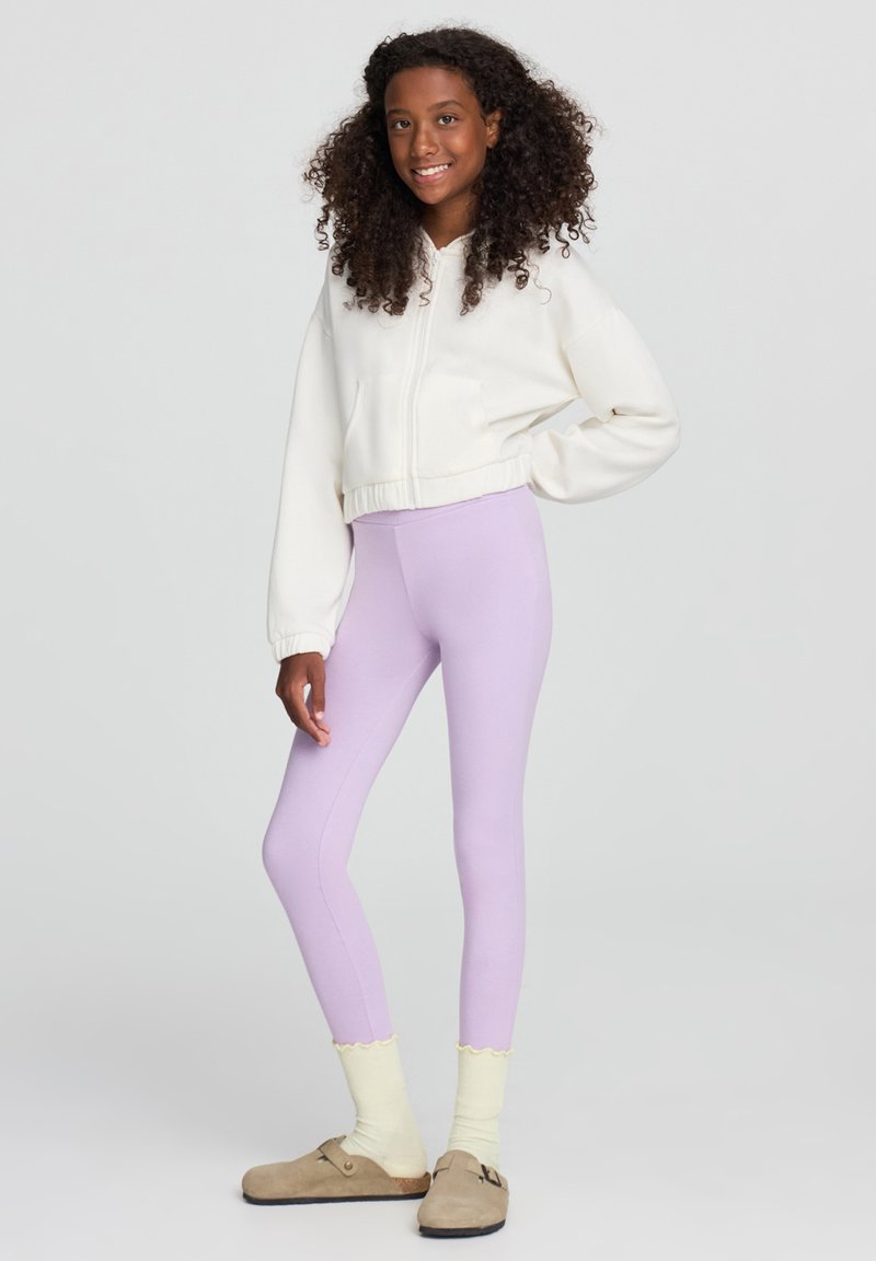 Leggings viola con una texture liscia, abbinati a un hoodie bianco corto e calzini giallo chiaro con bordo scallop, indossati con zoccoli beige.