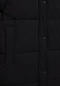 Veste matelassée noire avec une surface lisse, des coutures horizontales, des boutons-pression et une poche latérale pour le rangement.